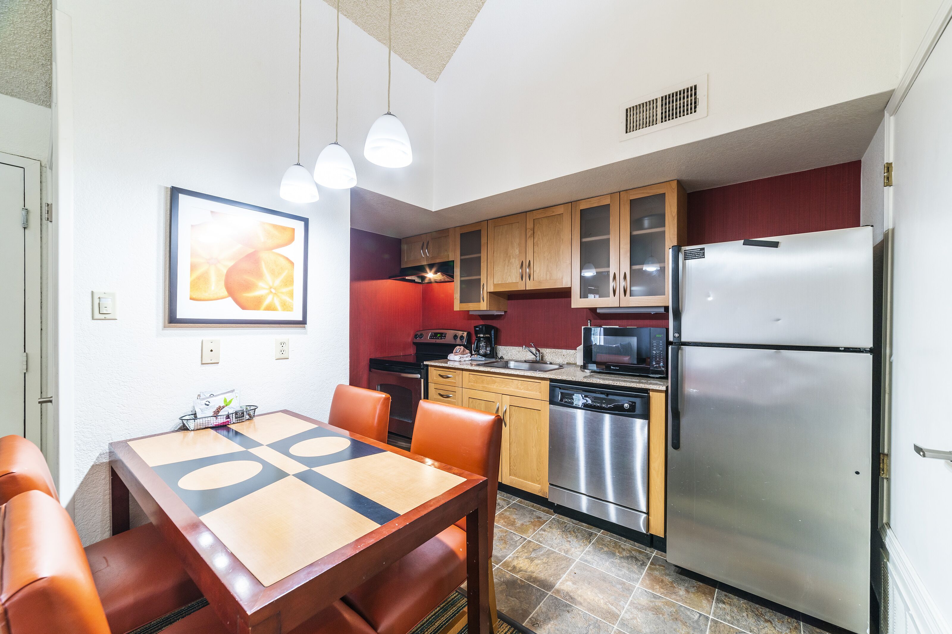 A compact kitchen with a dining area, featuring a stainless steel refrigerator, wooden cabinets, and a dining table with orange chairs.