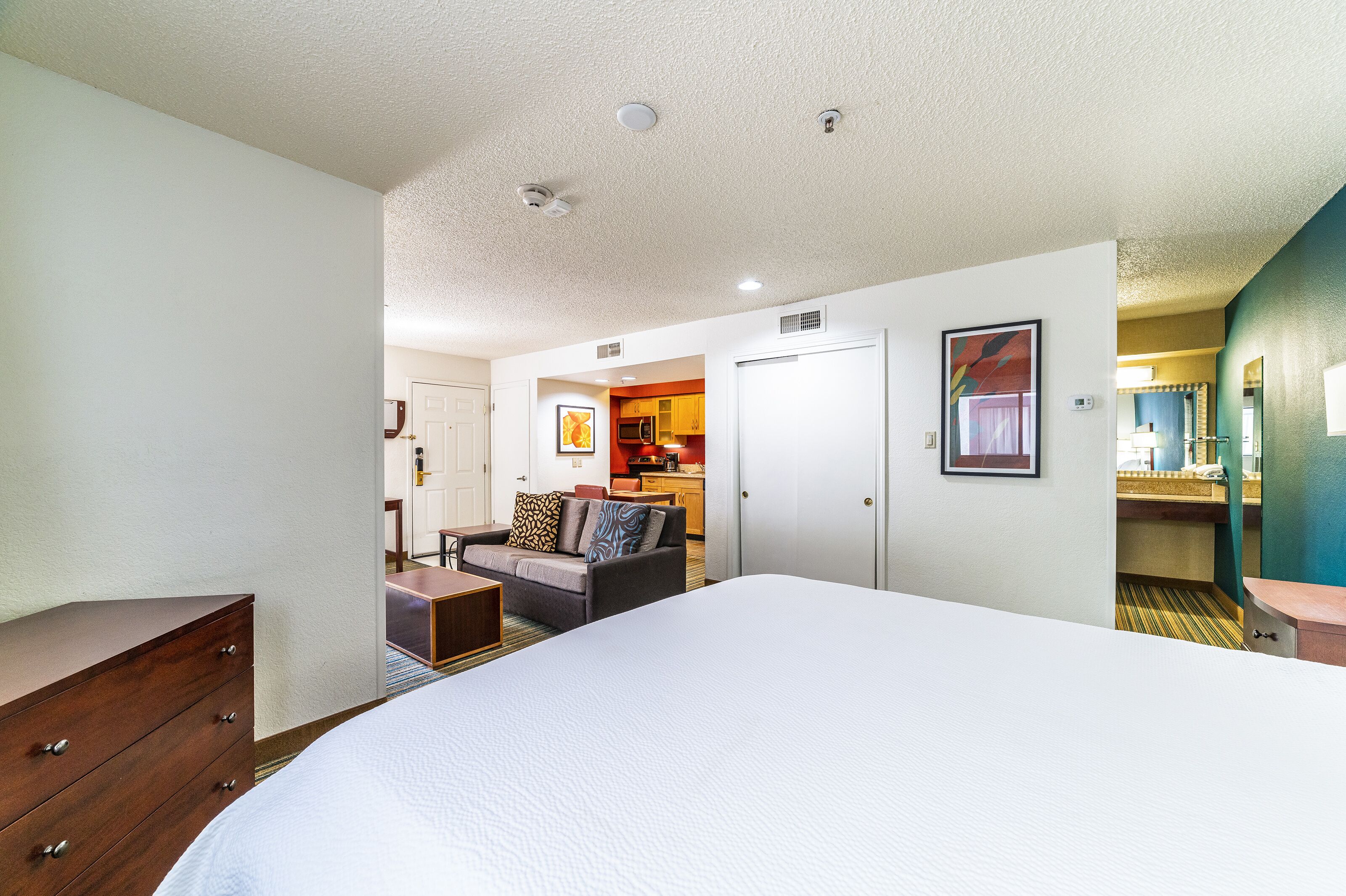 A hotel room with a large bed, a wooden dresser, a sofa, and a kitchenette in the background.