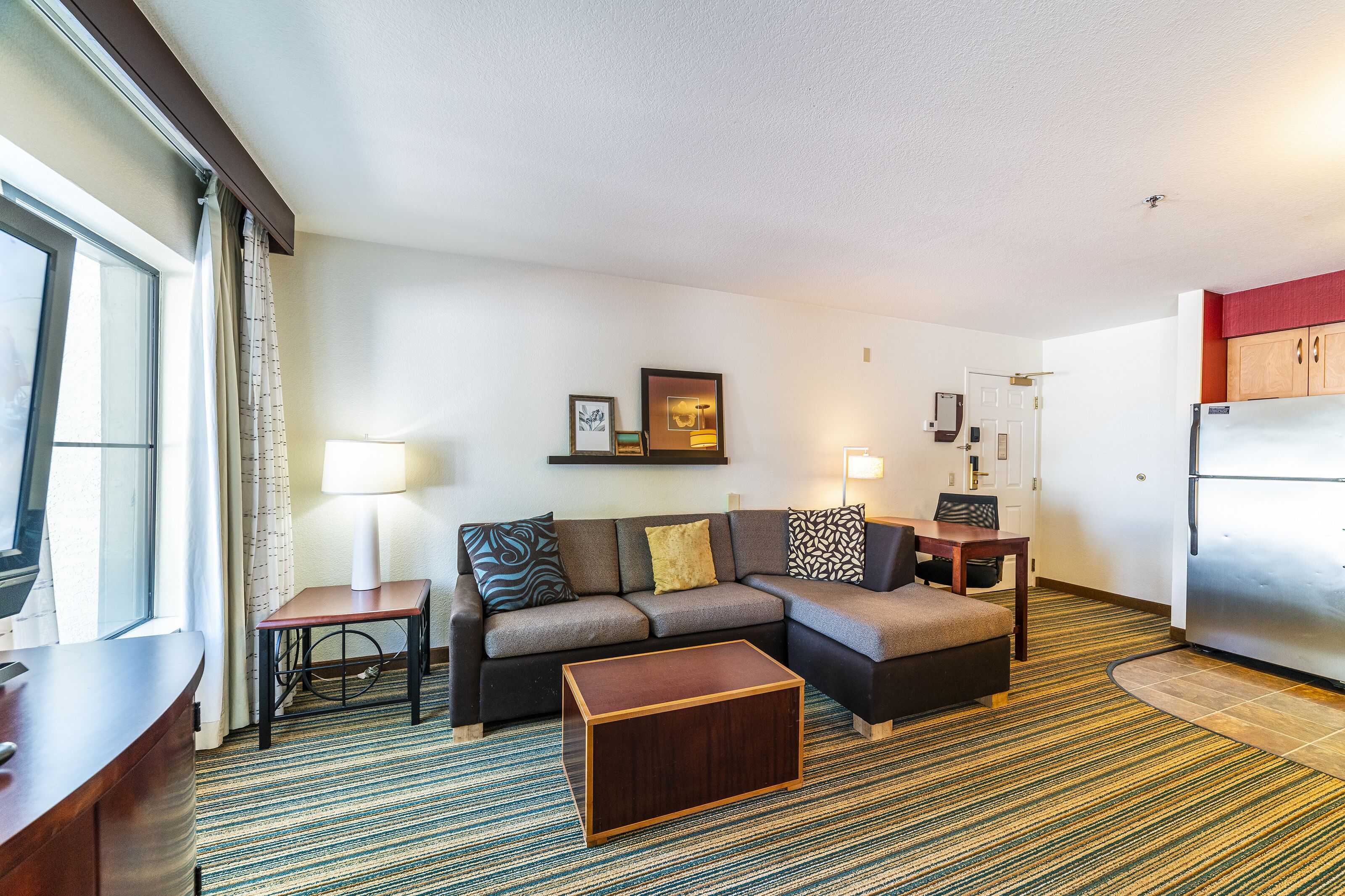 A compact hotel room with a sofa, a small table, a coffee table, a refrigerator, and a window with curtains.
