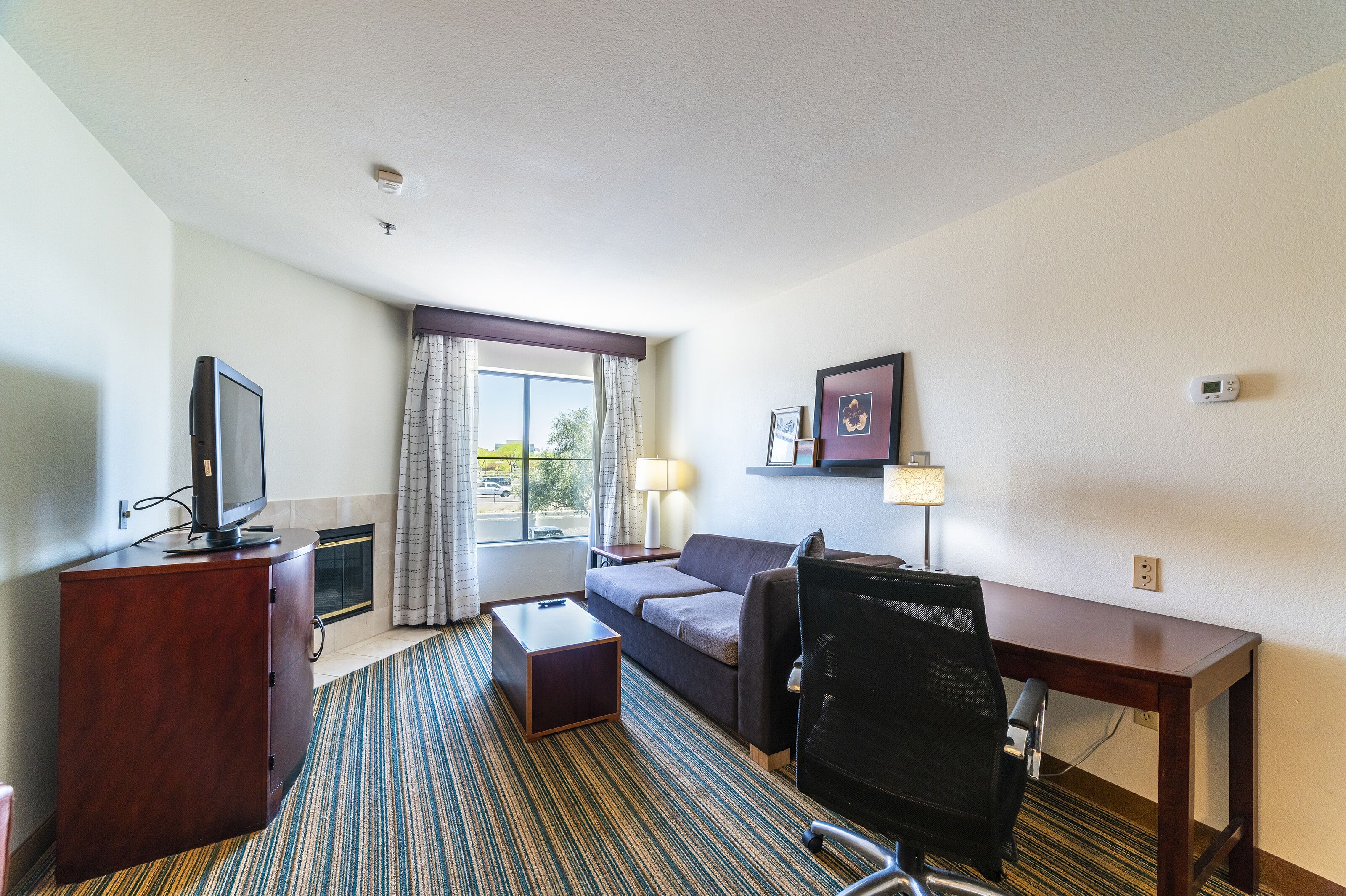 Ein Hotelzimmer mit Flachbildfernseher, einer Couch, einem Schreibtisch, einem Stuhl, einer Lampe und einem Fenster mit Vorhängen.