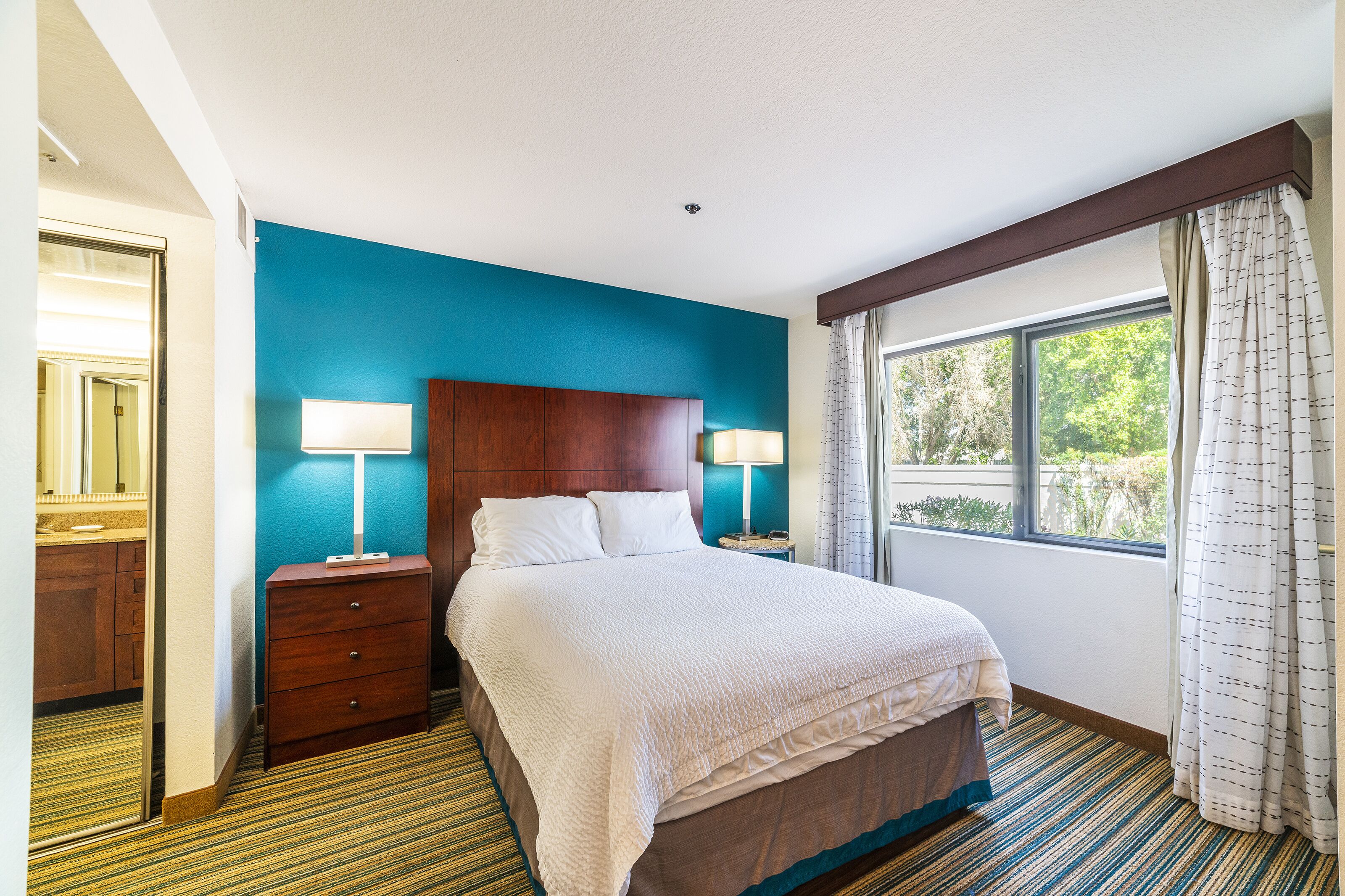 A hotel room with a large bed, two bedside tables with lamps, a wooden headboard, a window with curtains, and a view of greenery outside.