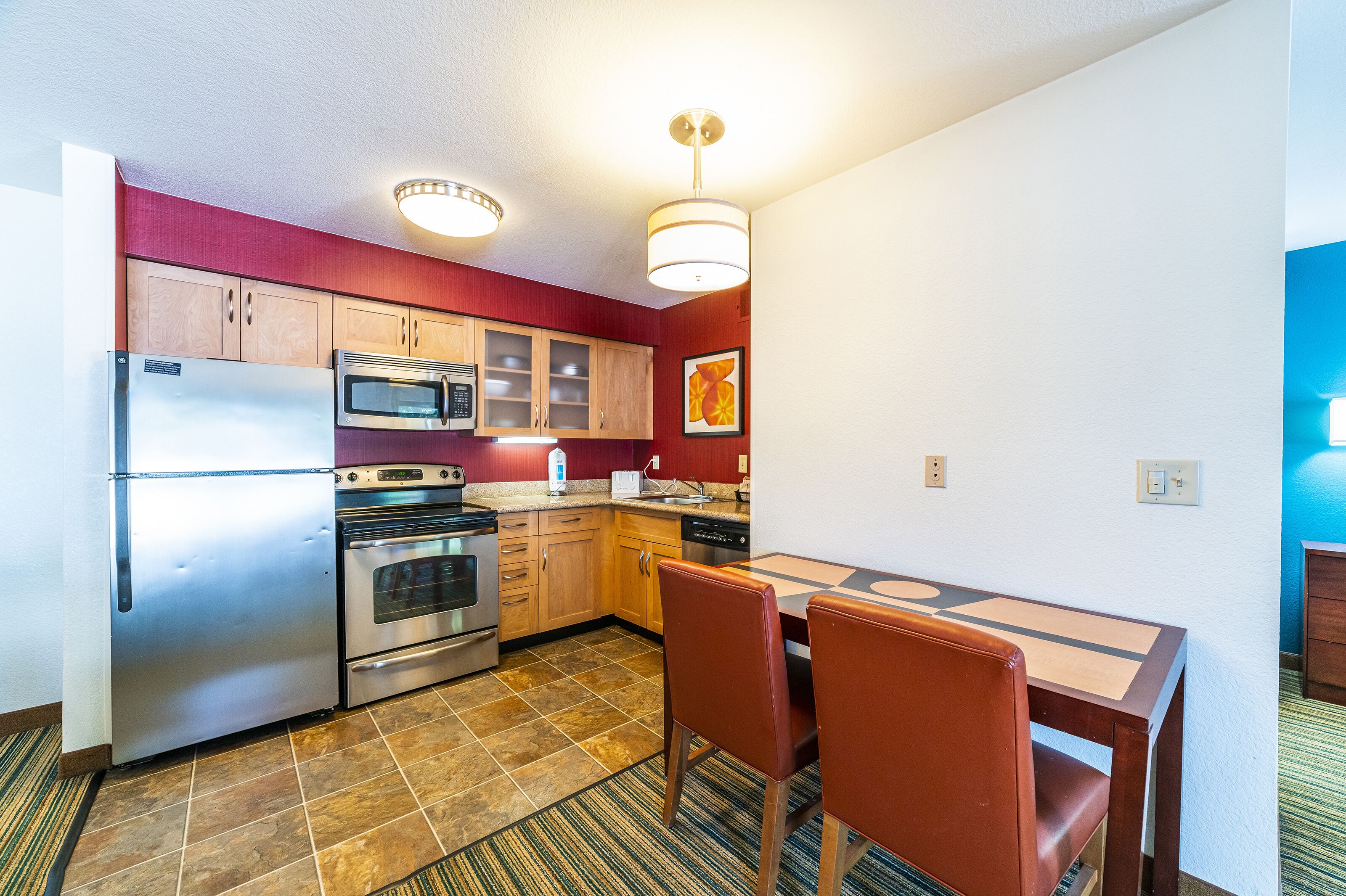 A compact kitchen with a refrigerator, microwave, stove, and dishwasher. There is a dining table with chairs and a patterned rug.