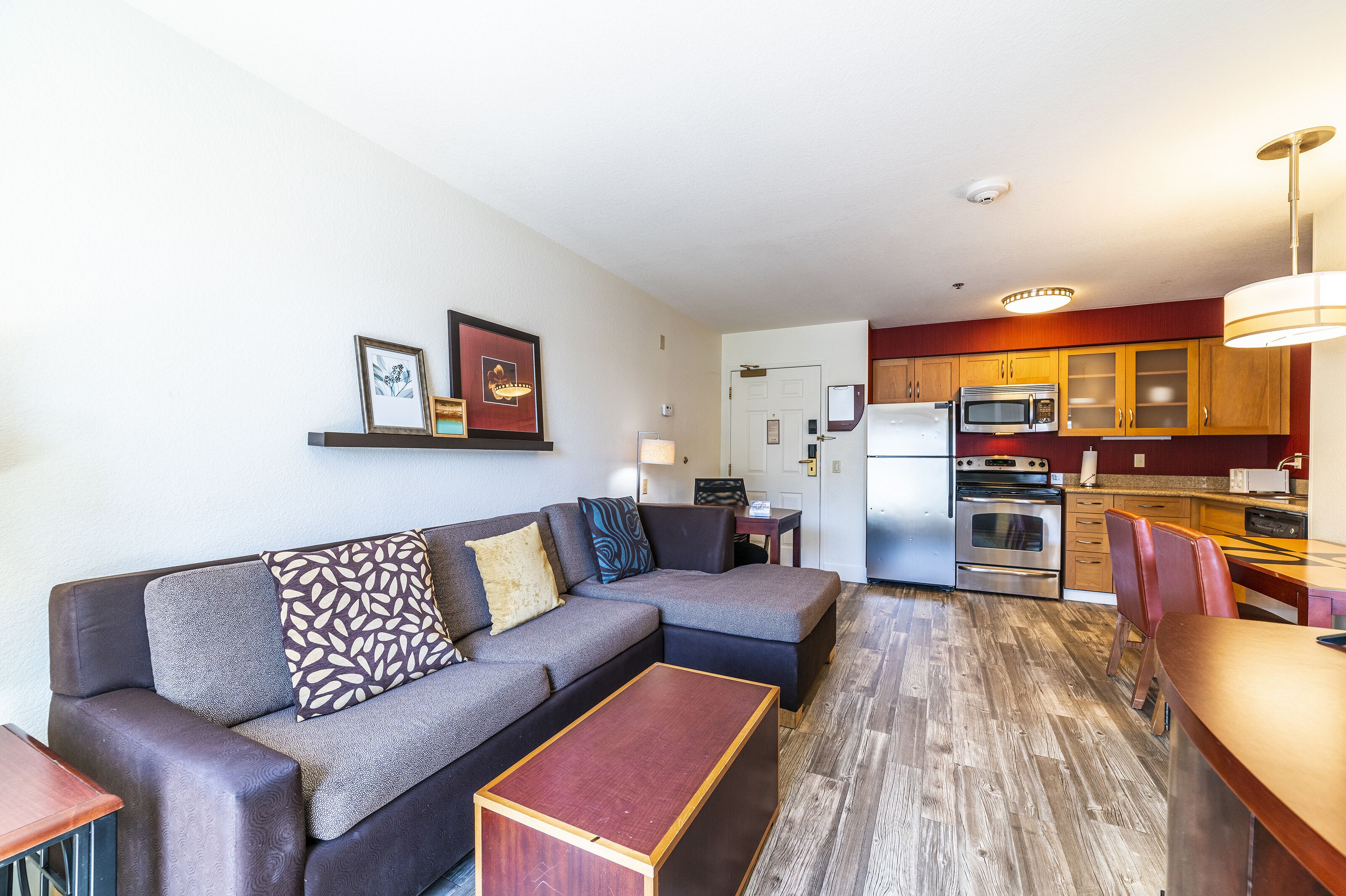 A modern living room with a sectional sofa, a wooden coffee table, and a kitchen area with wooden cabinets and stainless steel appliances.