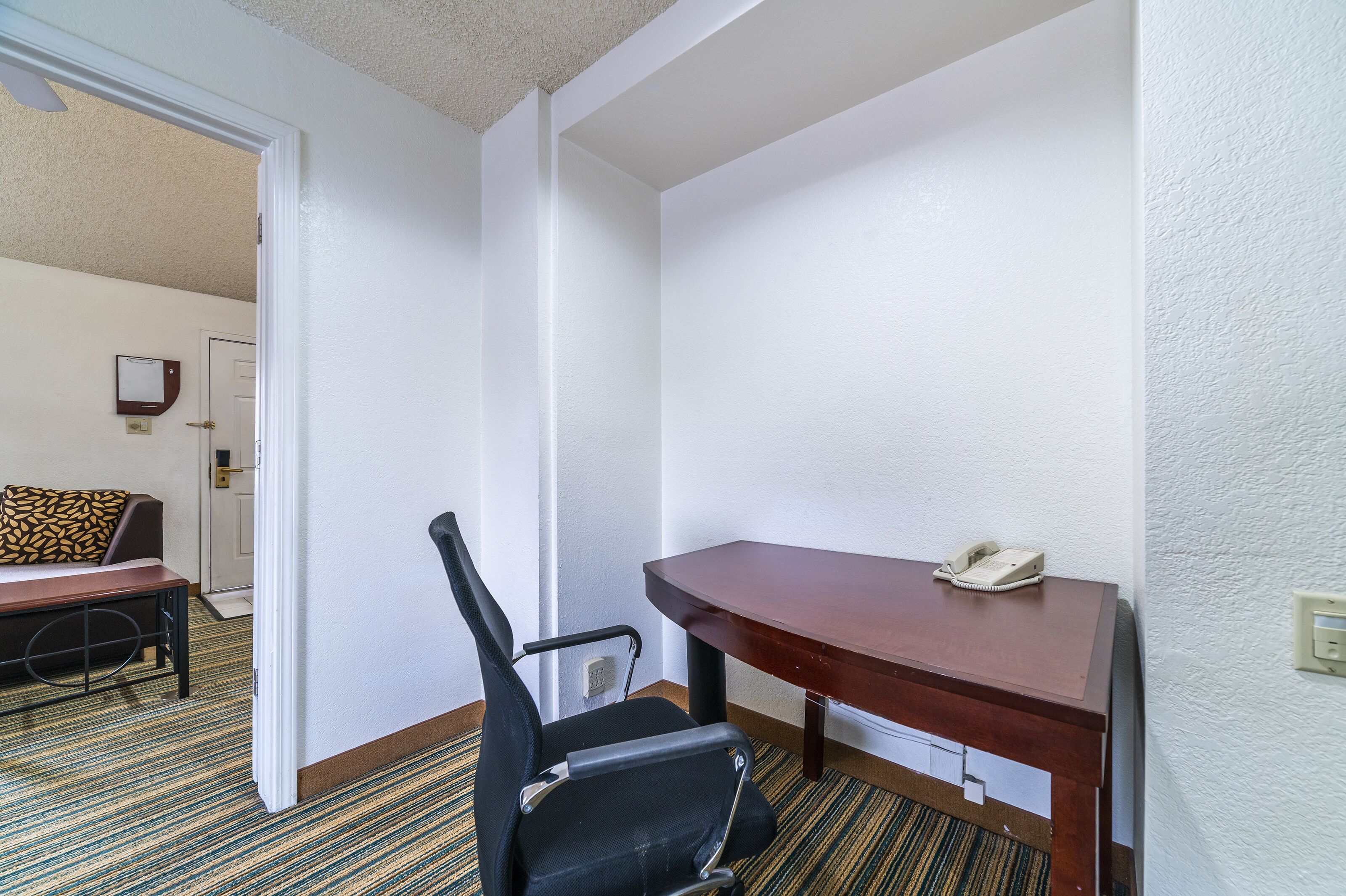 A hotel room with a wooden desk, a black office chair, a patterned sofa, and a door leading to another room.