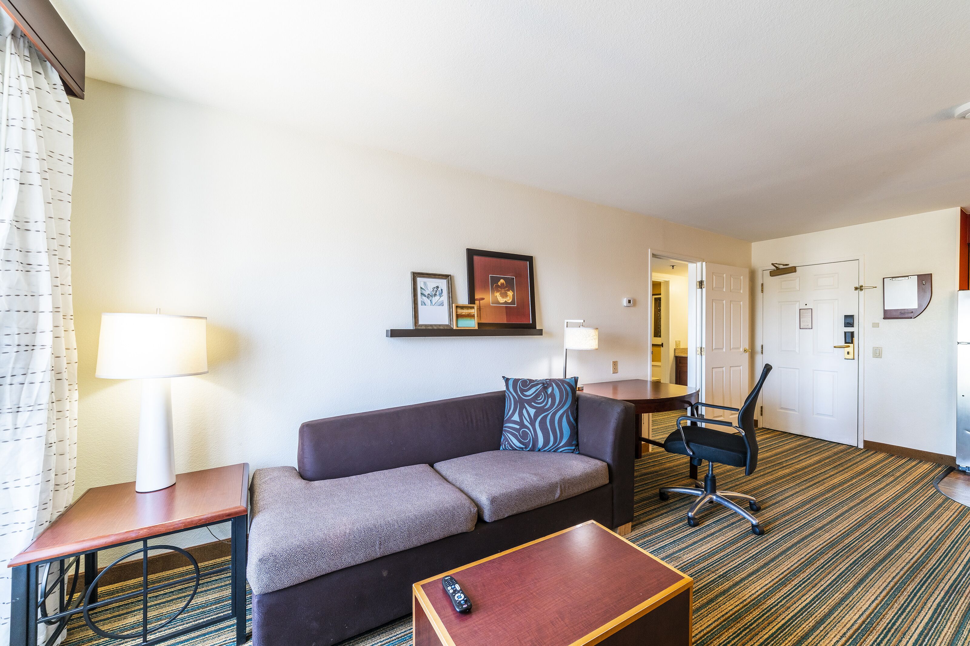 Une chambre d’hôtel avec un canapé, une petite table, une chaise, une lampe et une porte.