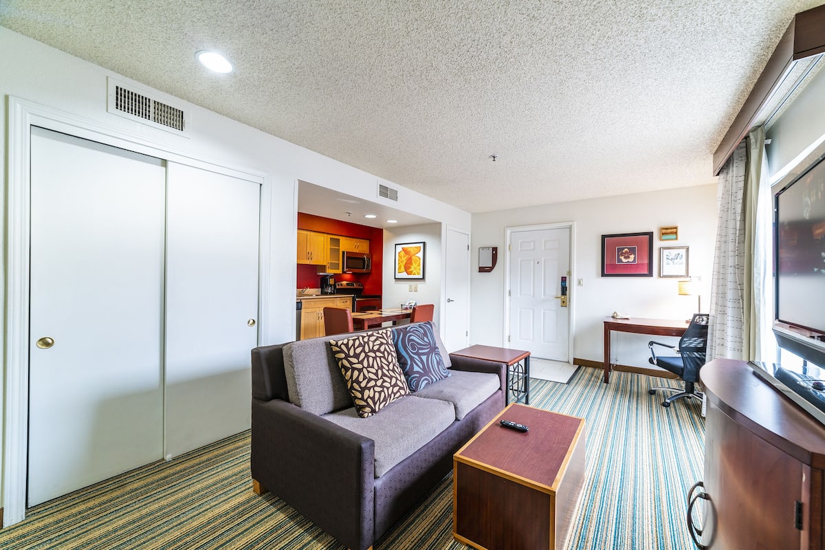 Quarto de hotel com sofá, mesa pequena, televisão, cozinha compacta e área de jantar.