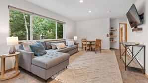 A modern living room with a grey sectional sofa, a dining table with chairs, a TV mounted on the wall, and a large window with a view of trees. - Luxury Home-Walk to everything in Frisco. Hot tub, ski, fireplace 3BD, 3.5BA



 (Frisco)