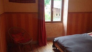 Una habitación con suelo de madera, una cama, una silla, un ventanal con cortinas y un tapiz en la pared.