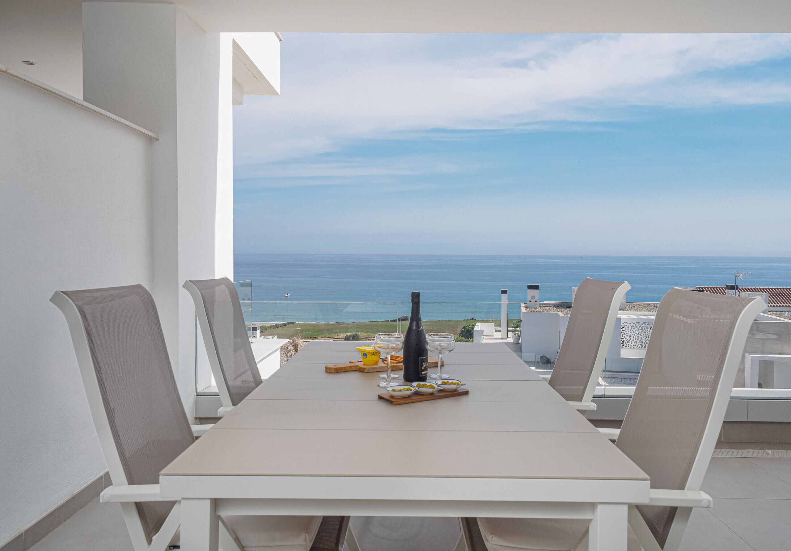 A table with four chairs, a bottle, and glasses on a balcony overlooking the sea.