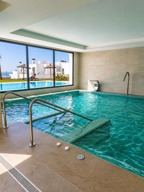 An indoor swimming pool with clear blue water, a metal ladder, and a view of a white building and palm trees outside. - The Links II Sea View 2473 La Alcaidesa Golf (La Alcaidesa)