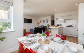 A modern kitchen with white cabinets, a dining area with a table set for four, and a living area with a sofa and a TV.