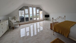 A bedroom with a sloped ceiling, a bed, a chair, a TV, and a large window with a view. - Littlestairs (Shanklin)