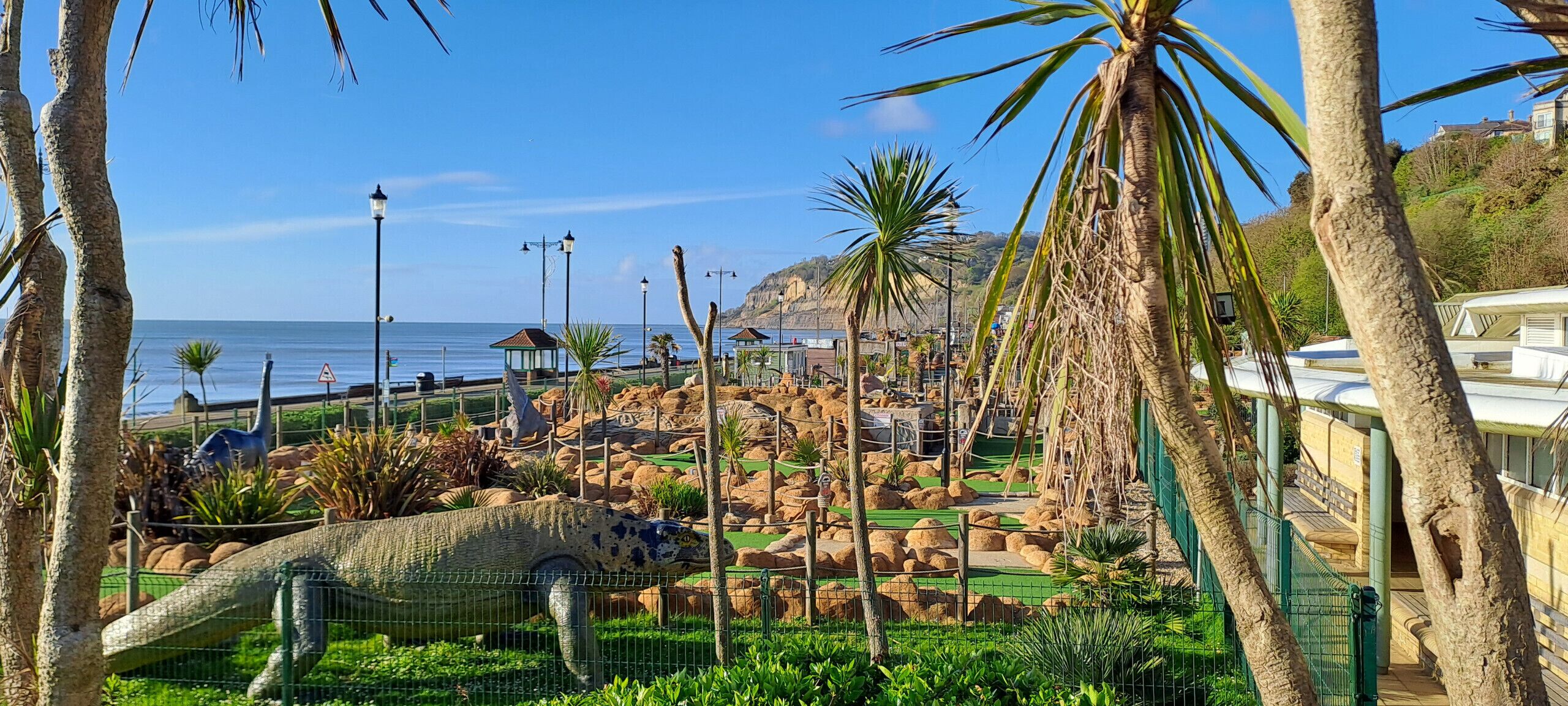 A dinosaur exhibit with a large model of a dinosaur in a fenced enclosure, surrounded by palm trees and rocks.