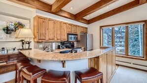 A kitchen with wooden cabinets, a large island with stools, a refrigerator, and a window with a view of trees. - Ski-in Condo w Vail Views near Gondola, TT5C (Vail)
