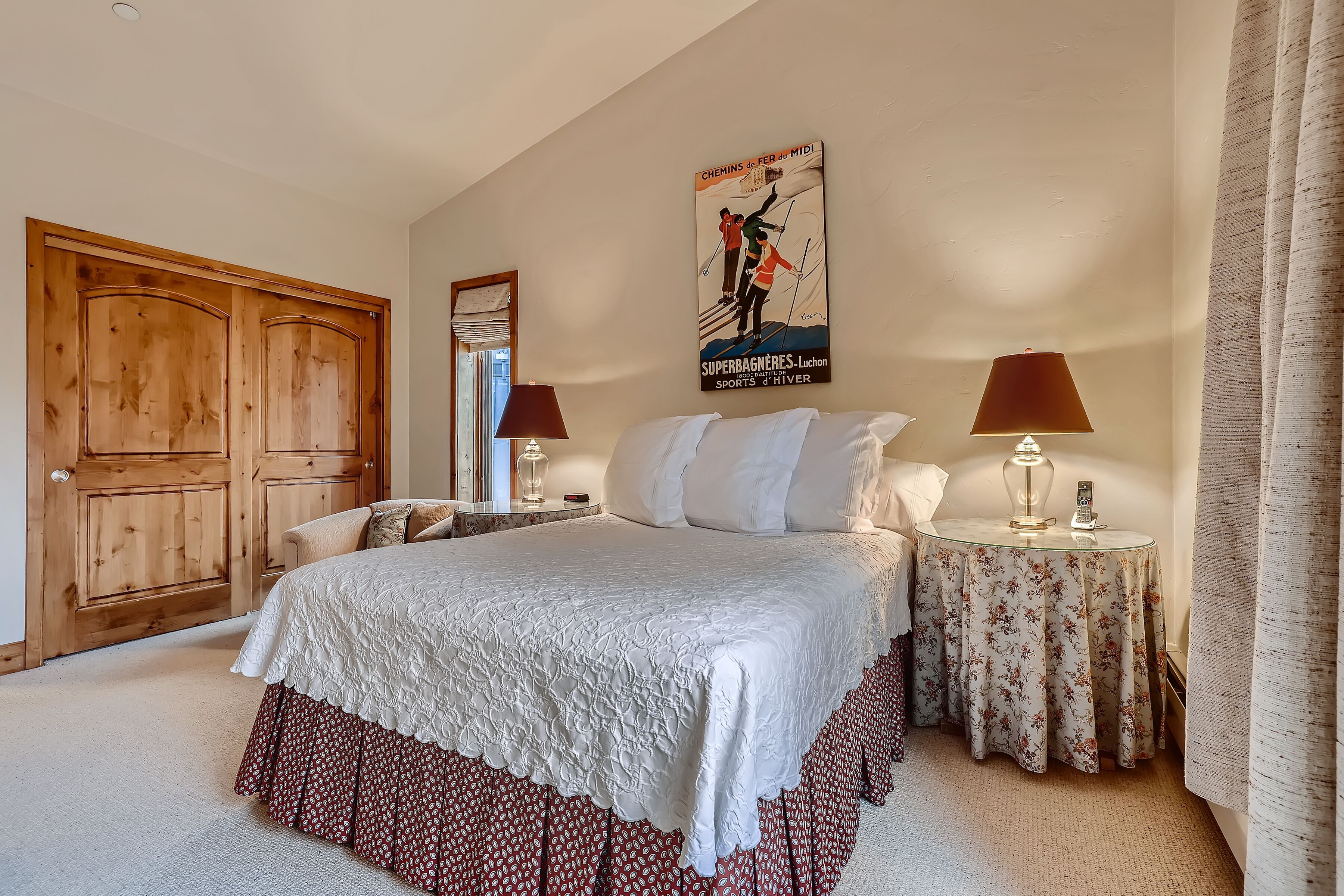 Une chambre avec un grand lit, une armoire en bois, deux tables de chevet avec des lampes, une affiche au mur et une fenêtre avec des rideaux.