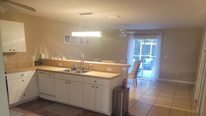 Une cuisine avec des armoires blanches, un évier et un plan de travail. On y trouve un ventilateur de plafond, une fenêtre avec des rideaux et une porte donnant sur un patio.