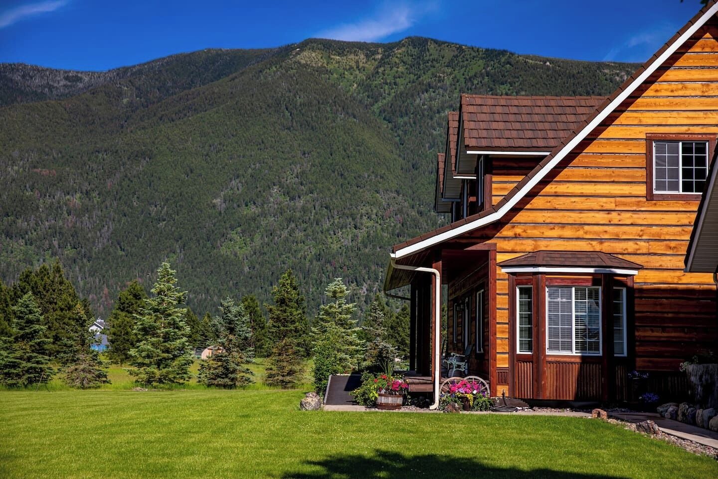 Une cabane en rondins à deux étages, dotée d’une véranda qui fait le tour de la maison, et située au pied d’une montagne boisée.
