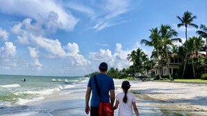 Un couple se promène sur une plage, avec des vagues, des palmiers et une maison en arrière-plan.