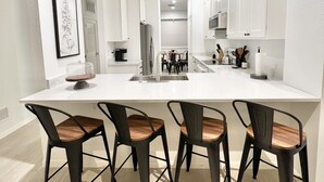 Une cuisine moderne avec des meubles blancs, un grand îlot central doté de tabourets de bar et une reproduction botanique encadrée accrochée au mur.