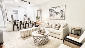 Un salon moderne avec un canapé blanc, une table basse ronde et un coin repas comprenant des chaises, avec une cuisine en arrière-plan.
