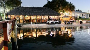 Un restaurant doté d’un toit de chaume, de tables en plein air et d’un élément décoratif aquatique en premier plan.