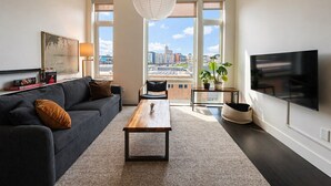 Una sala de estar moderna con un sofá, una mesa de centro, un televisor y ventanas amplias con vistas a la ciudad.