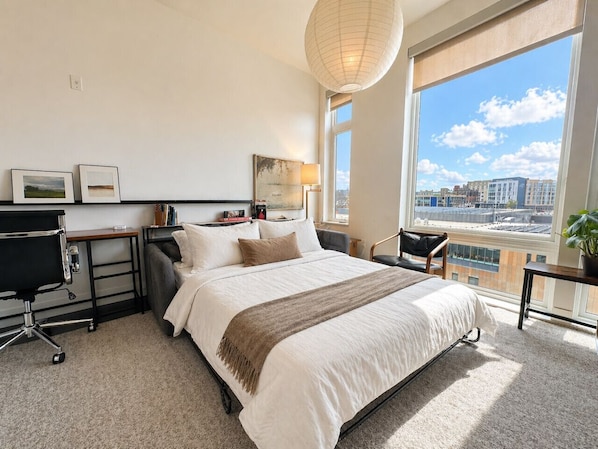 A modern hotel room with a large bed, a desk, a chair, a lamp, and a view of the city.