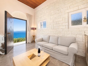 A modern living room with a stone wall, a sofa, a coffee table with a bottle and glasses, and a view of the sea.