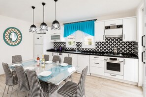 Uma cozinha moderna com armários brancos, painel de backsplash com padrão preto e branco, e um espaço de jantar com cadeiras xadrez e uma mesa de vidro.