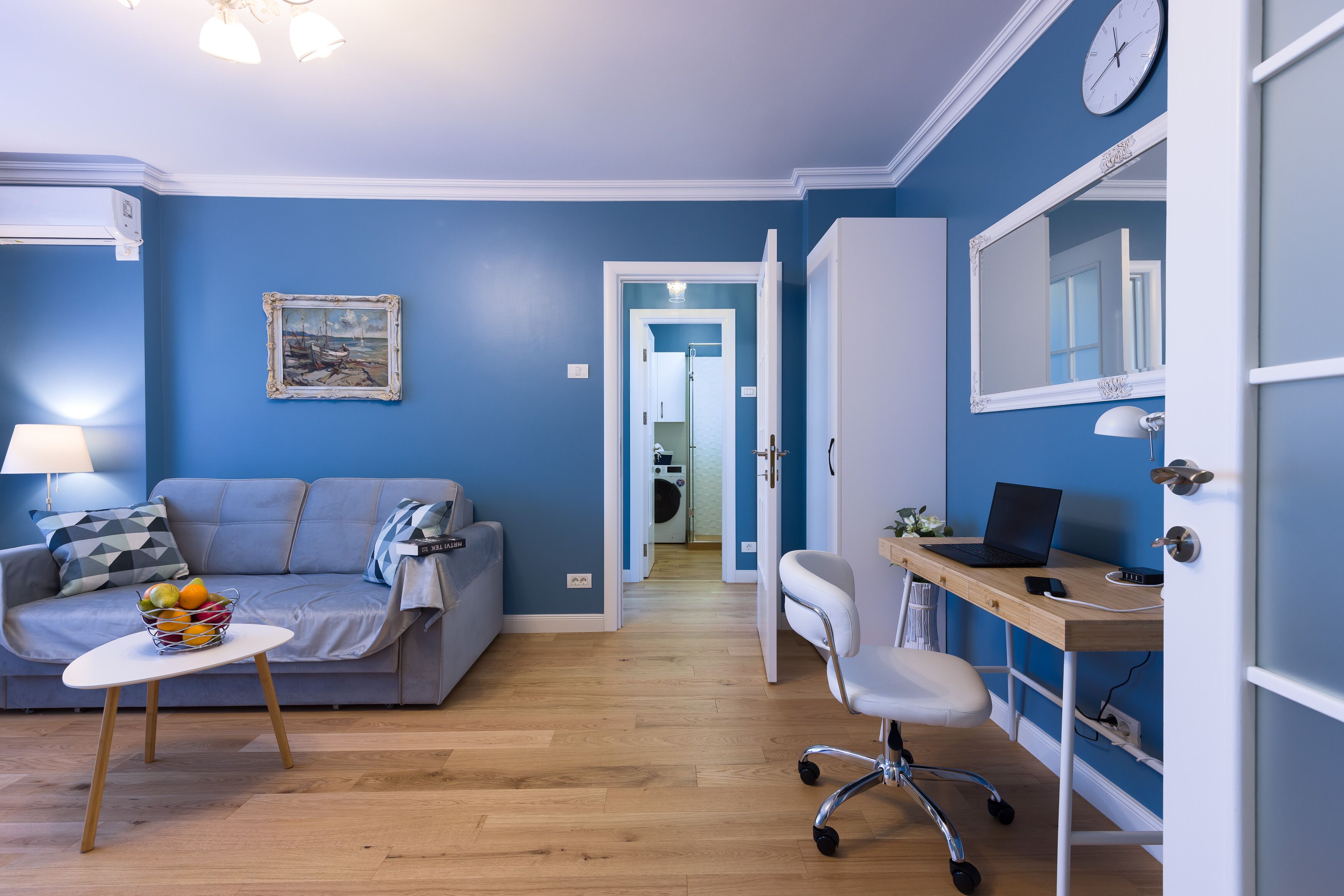 A living room with a blue wall, a sofa, a coffee table with fruit, a desk with a laptop, and a mirror.