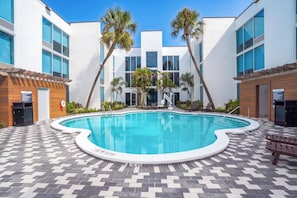 Una piscina con solárium de baldosas, rodeada de palmeras y edificios modernos.