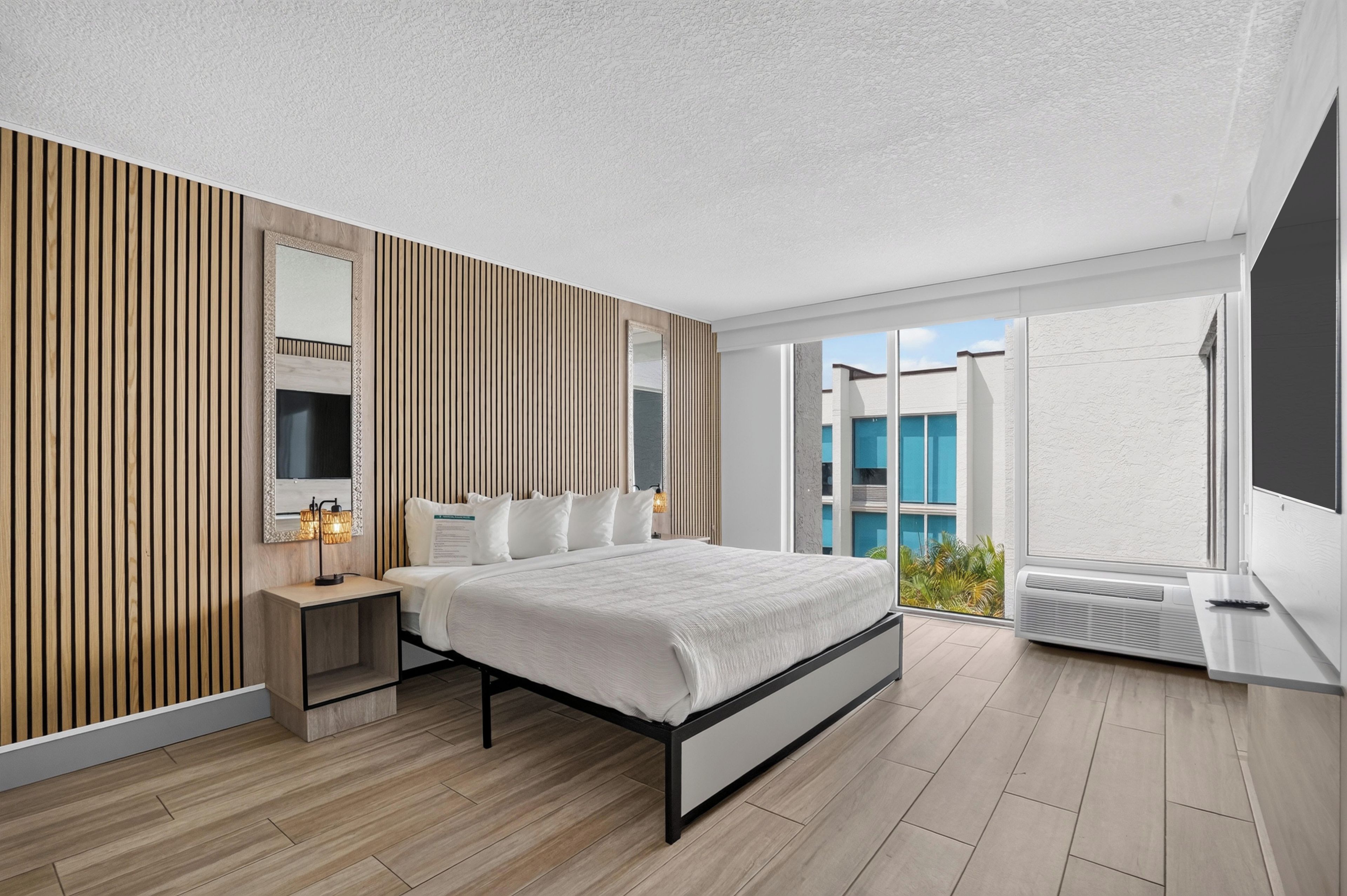 A modern bedroom with a bed, bedside tables, a TV, and a large window with a view of buildings.