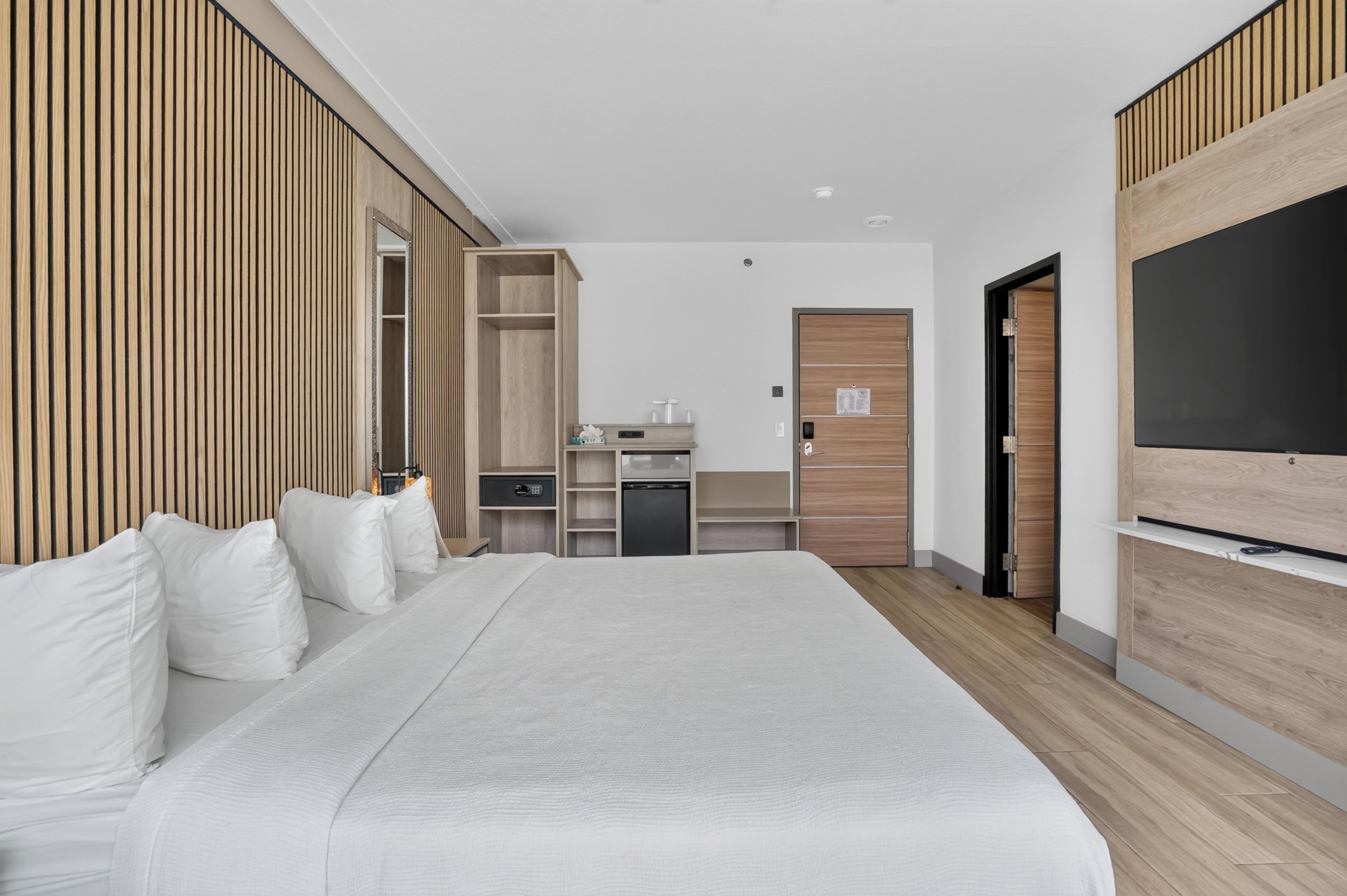 A modern hotel room with a large bed, a TV, a kitchenette, and wooden paneling on the walls.