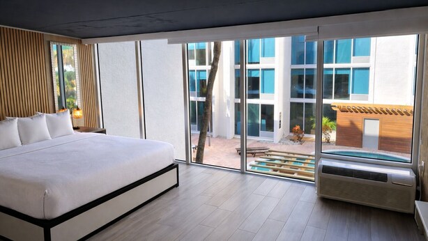 Une chambre d’hôtel moderne dotée d’un grand lit, de portes-fenêtres coulissantes donnant sur un espace extérieur et offrant une vue sur un immeuble aux fenêtres bleues.