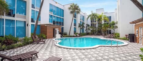 A swimming pool surrounded by lounge chairs and a checkered tile floor, with palm trees and a building in the background.