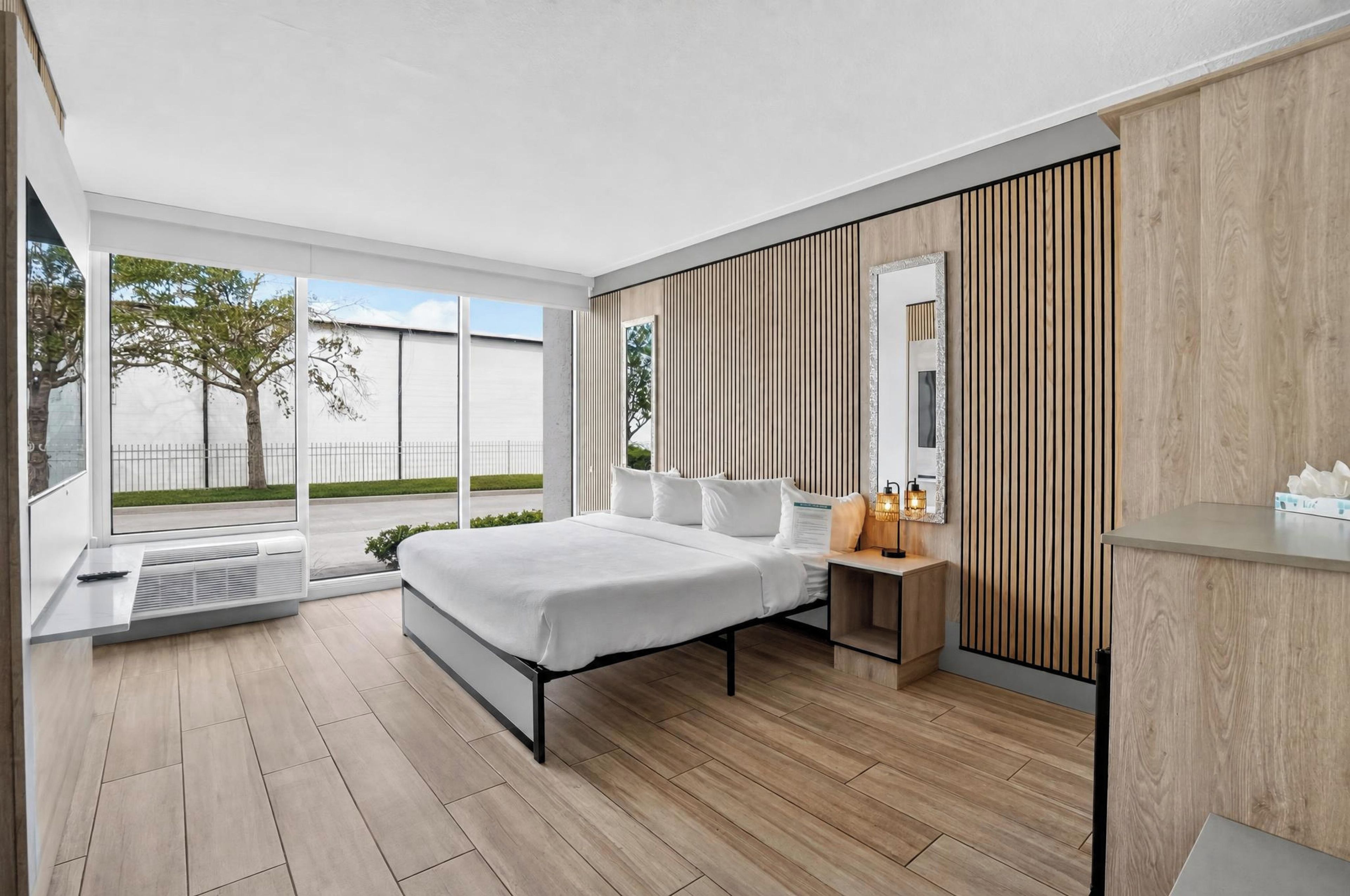 A modern bedroom with a large bed, a bedside table with a lamp, a mirror, and a view of the outdoors through large windows.