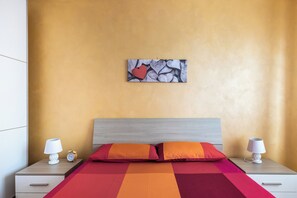 Un dormitorio ordenado con cama, almohadas, mesita de noche, lámpara y una obra de arte en la pared.