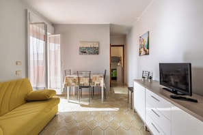 A living room with a yellow sofa, a dining table with chairs, a TV, and a kitchen area.