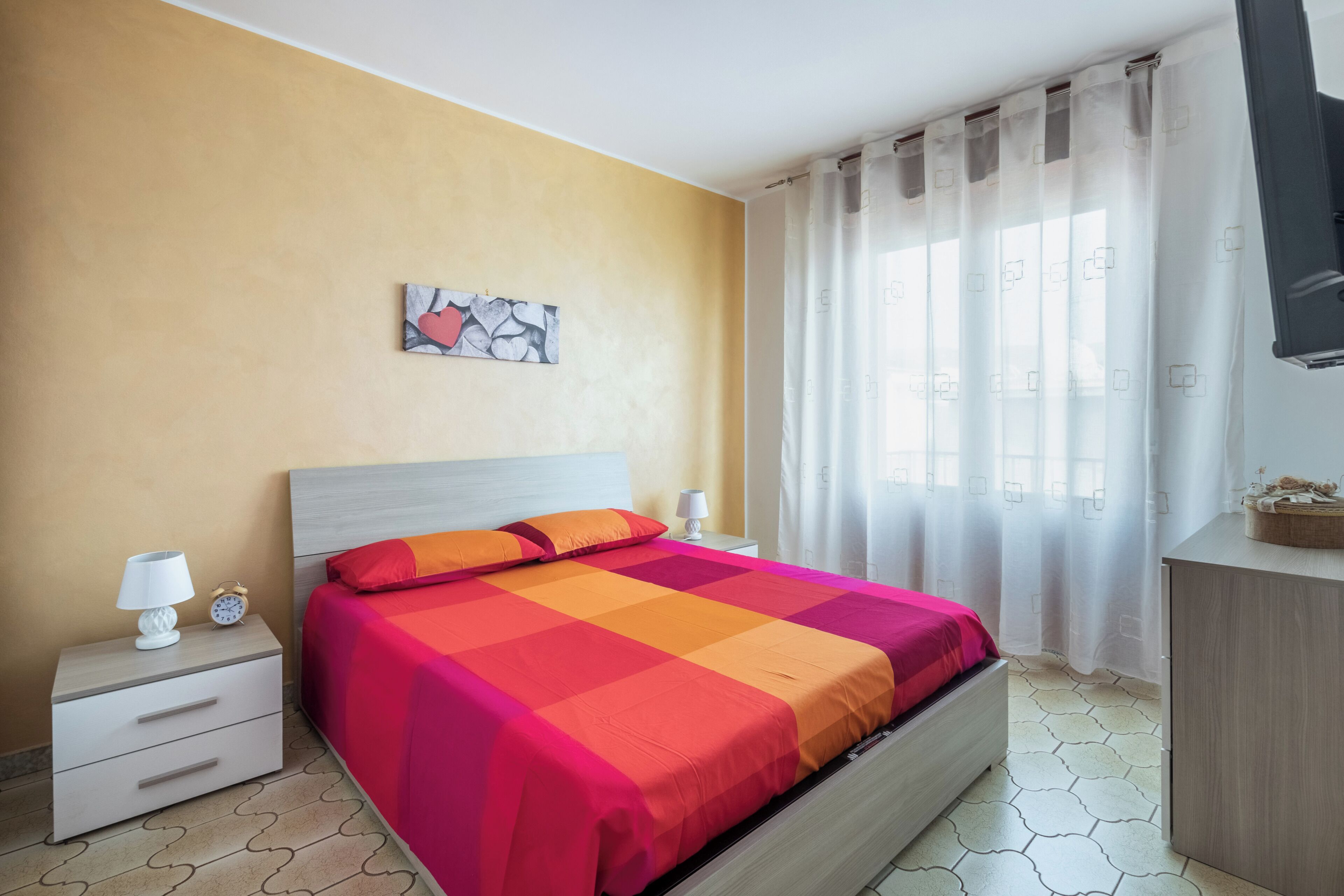 Una camera da letto con un letto, comodini, una sveglia a muro, un quadro e una finestra con tende.