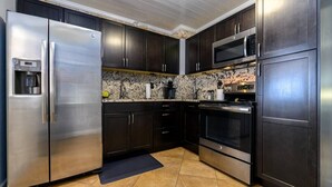 A modern kitchen with dark cabinetry, stainless steel appliances, and a granite backsplash. - Cozy Well-Equipped Home In The Heart Of South Tampa. Amazing Fenced-in Backyard! (Tampa)