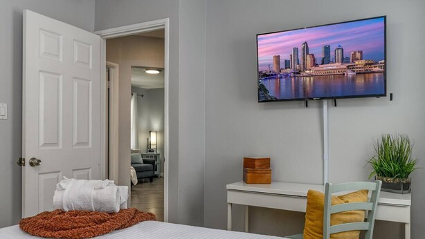 A hotel room with a bed, a desk, a TV displaying a cityscape, and a door leading to another room. - Cozy Well-Equipped Home In The Heart Of South Tampa. Amazing Fenced-in Backyard! (Tampa)