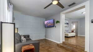 A modern living room with a grey sofa, a wooden coffee table, and a mounted TV. The room has a tiled floor and a ceiling fan. - Cozy Well-Equipped Home In The Heart Of South Tampa. Amazing Fenced-in Backyard! (Tampa)