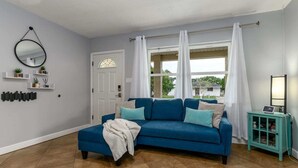 A living room with a blue sofa, a teal side table, a white door, and a window with white curtains. - Cozy Well-Equipped Home In The Heart Of South Tampa. Amazing Fenced-in Backyard! (Tampa)