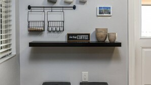 A kitchen with a coffee station, two wicker stools, and a shelf with a coffee sign. - Cozy Well-Equipped Home In The Heart Of South Tampa. Amazing Fenced-in Backyard! (Tampa)