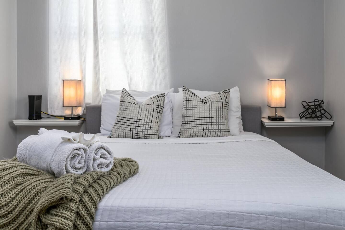 A neatly made bed with white linens, two checkered pillows, and a rolled towel on a white bedspread. There are two bedside tables with lamps and a small shelf with a black object.