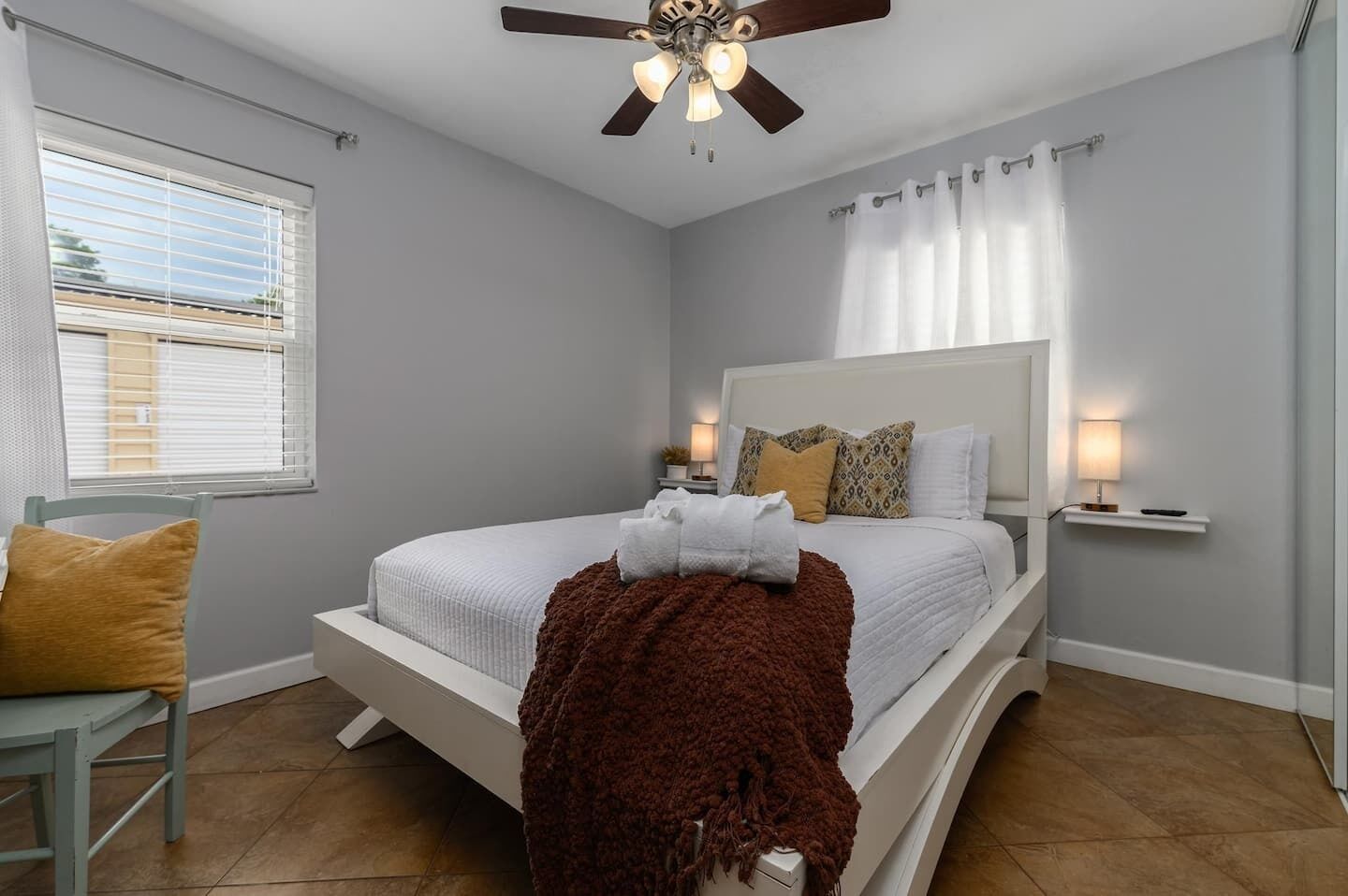 A bedroom with a bed, a ceiling fan, a window with blinds, and a bedside table with a lamp.