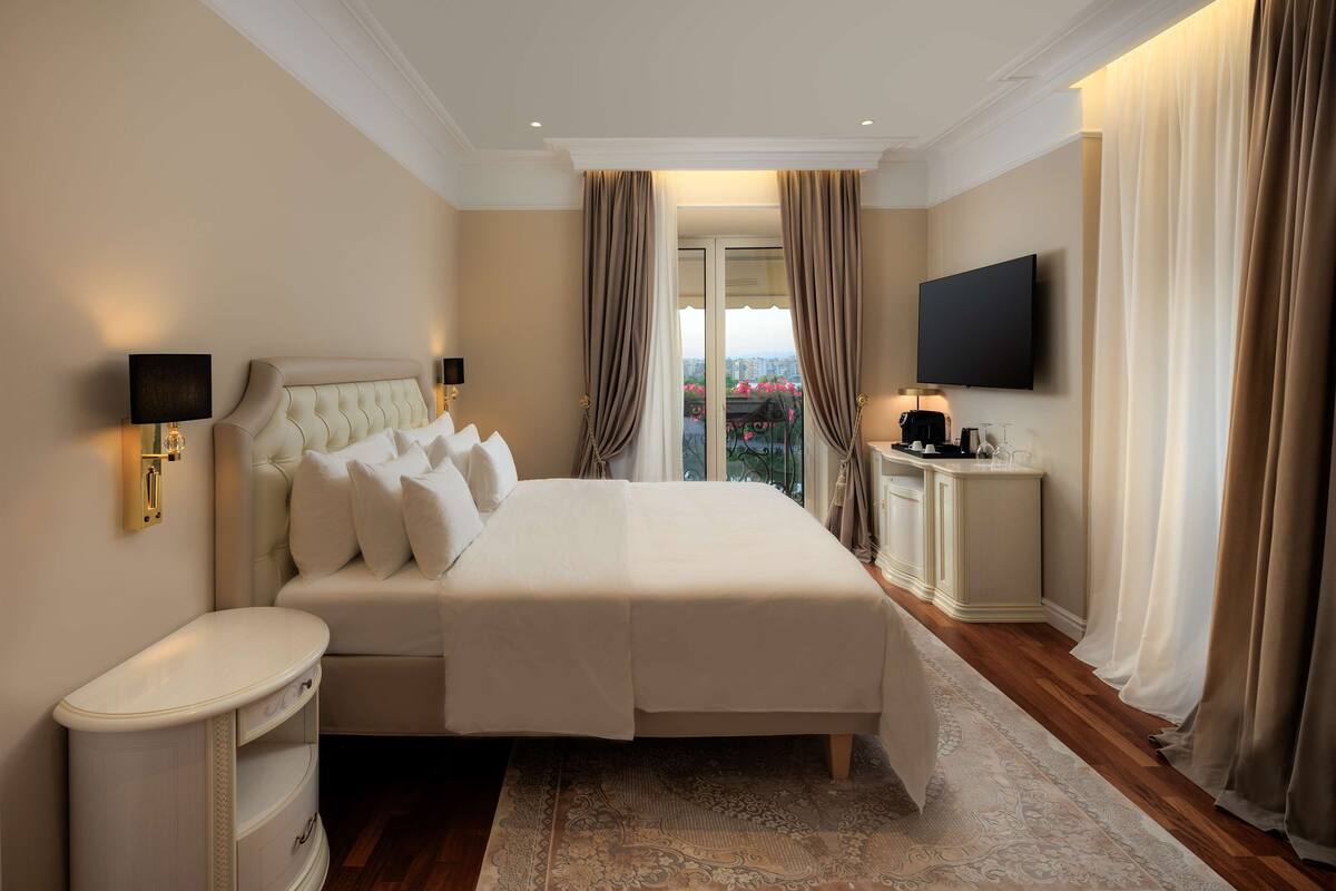 Habitación de hotel con una cama grande, mesitas de noche, un televisor de pantalla plana y vistas a la ciudad desde la ventana.
