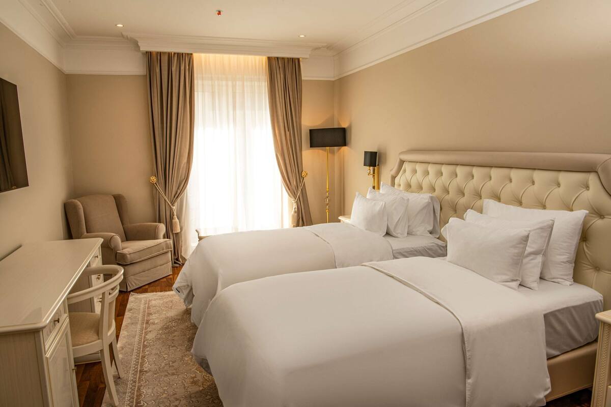 Habitación de hotel con dos camas, un escritorio, una silla y un ventanal con cortinas.