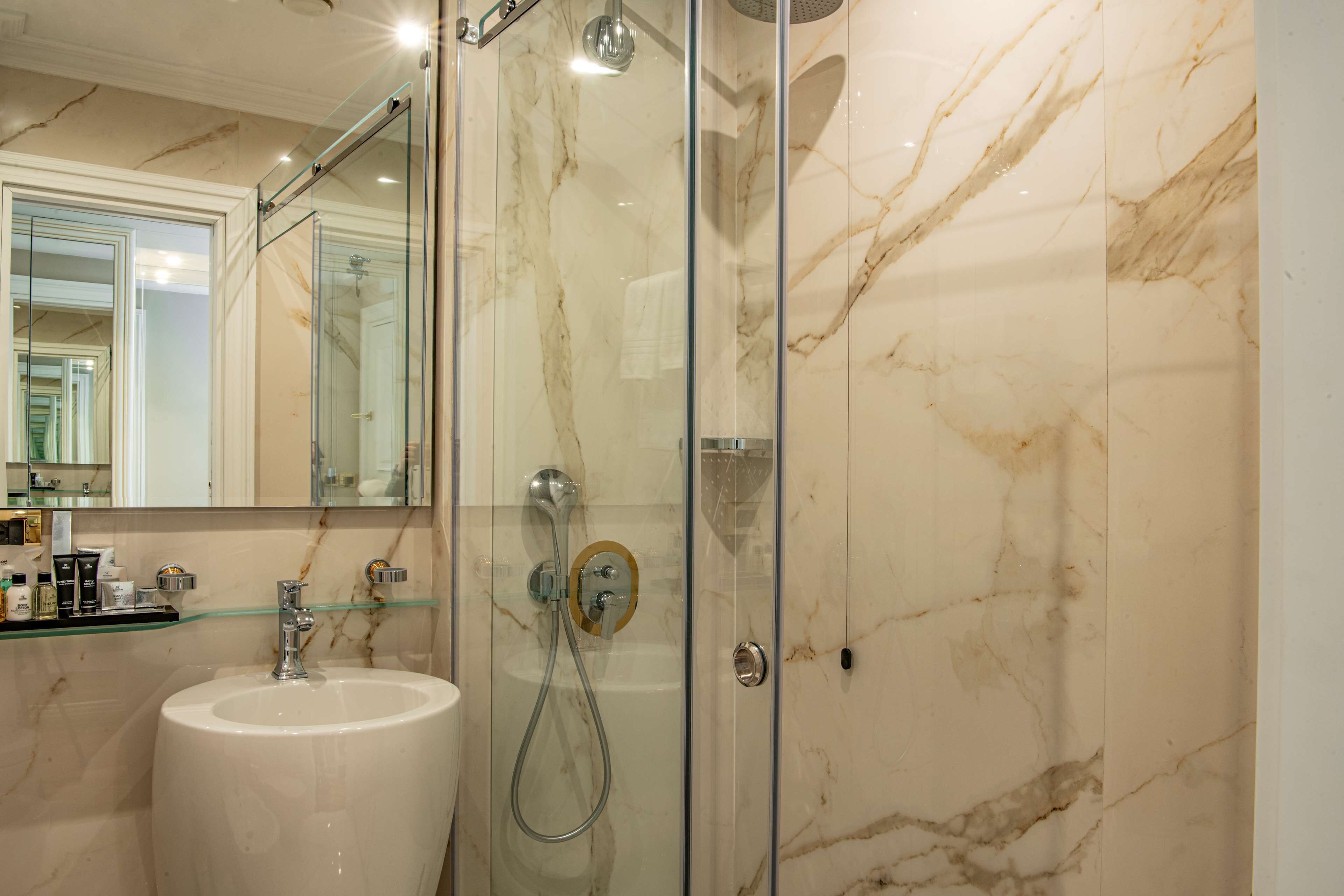 Une salle de bain dotée d’une douche à parois de verre, d’un lavabo en marbre et d’une étagère en verre où sont rangés les articles de toilette.
