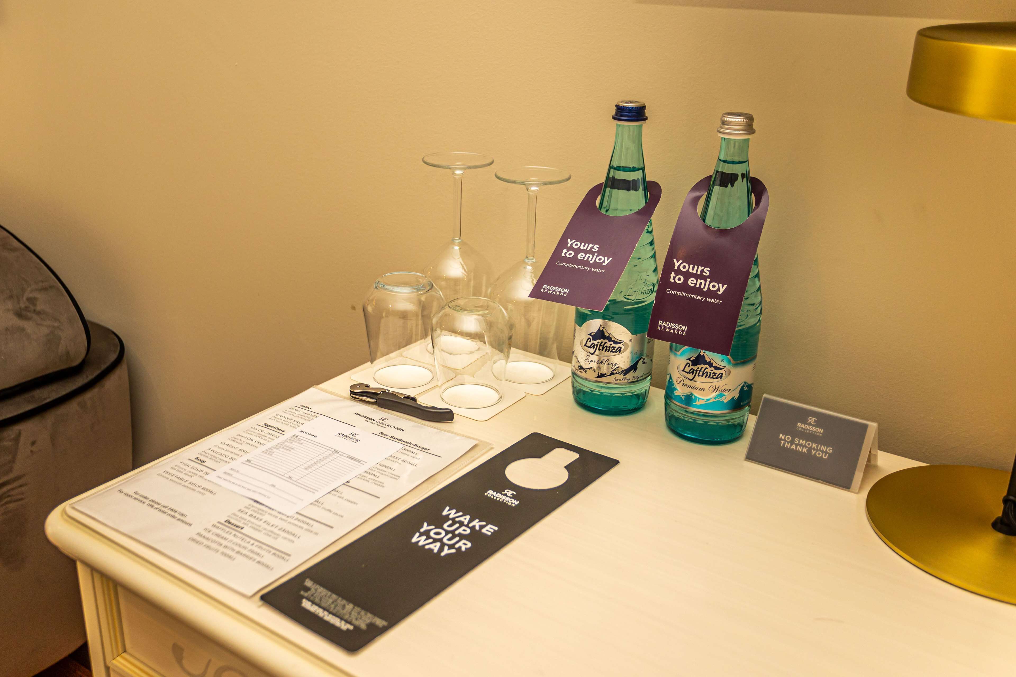 A hotel room with a minibar, a menu, a wake-up sign, and two bottles of water labeled 'Yours to enjoy.'